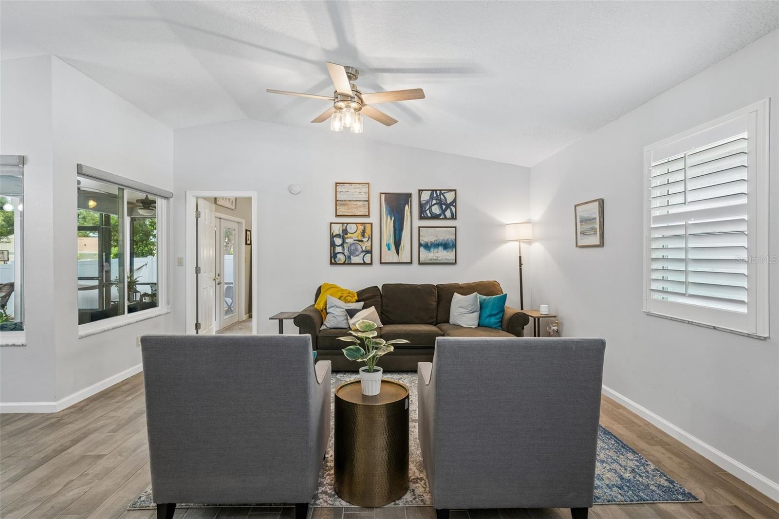 Living room with vaulted ceilings, wood-look tile floors, plantation shutters, and views out to the patio.