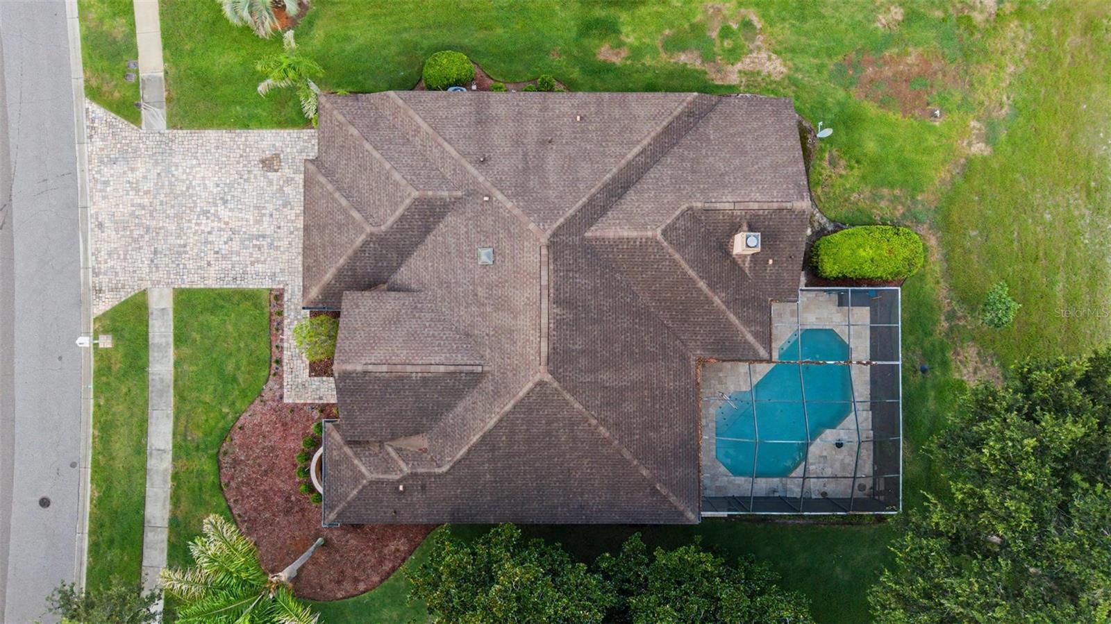 Drone aerial view directly above the home, showcasing the roofline, backyard pool, and surrounding lot. The image highlights the paver driveway, mature landscaping, and overall property layout, offering a clear perspective of the home’s footprint and outdoor living areas.