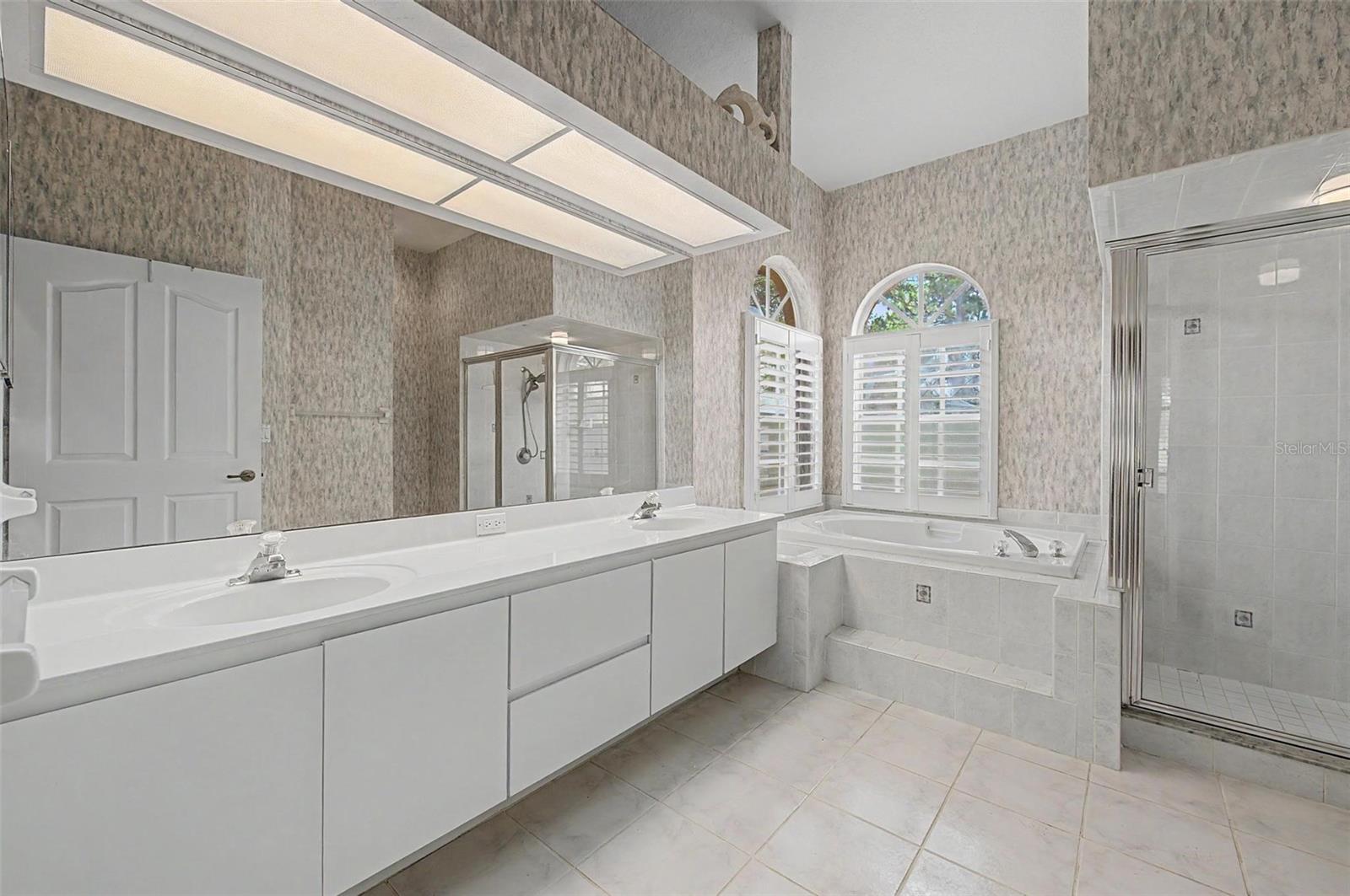 Master bathroom featuring a spacious five-piece layout with an extended dual-sink vanity. The bathroom includes a soaking tub positioned beneath two windows with plantation shutters, providing abundant natural light. A separate walk-in shower and a private toilet area, partially enclosed by a privacy wall, complete this well-designed and functional space.