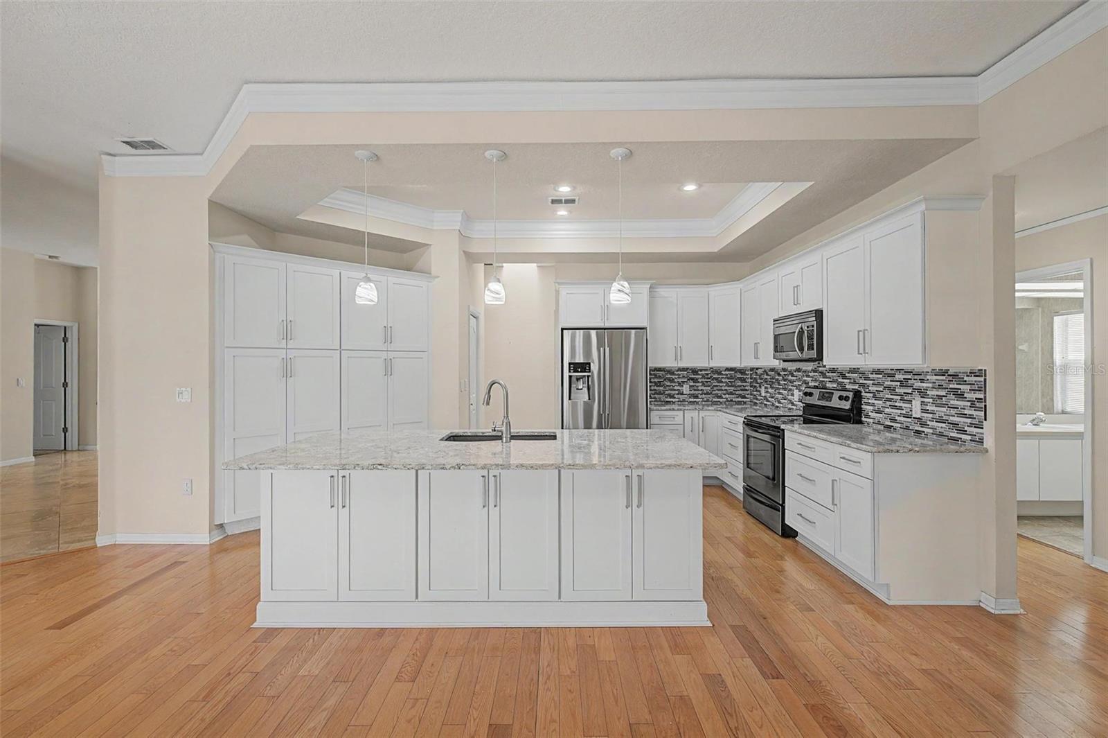 Beautifully updated kitchen featuring a bright, open layout with high ceilings and detailed crown molding that enhances the overall space. The kitchen offers an abundance of white cabinetry for ample storage, a large center island with granite countertops, and stylish pendant lighting. A modern tile backsplash extends throughout the cooking area, complementing the stainless steel appliances, including a refrigerator, range, microwave, and dishwasher. The wood-look flooring adds warmth, while the open design creates a seamless flow ideal for both everyday living and entertaining.