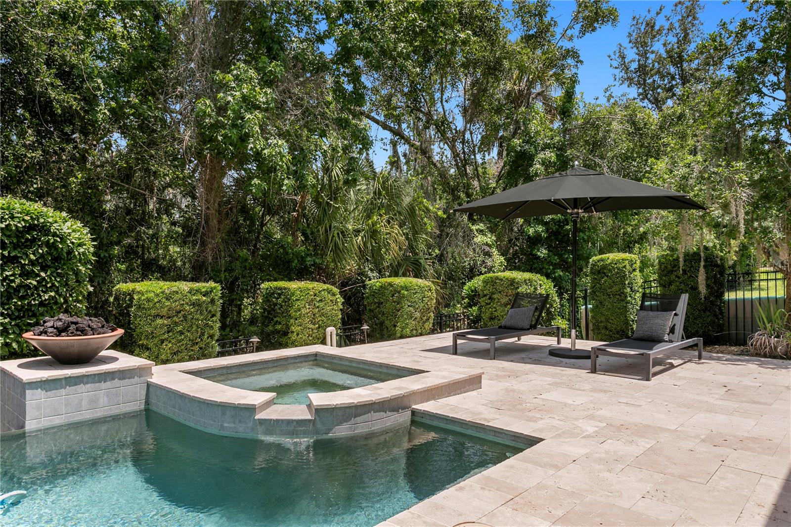 Custom geometric spa with a spilling waterfall feature, nestled within a travertine patio and framed by manicured hedges and lush conservation views.