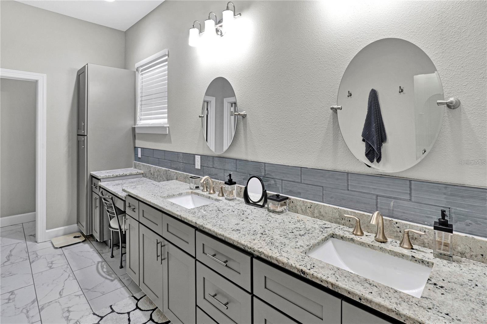 The luxurious primary bathroom features an extended double vanity with granite tops, a dedicated makeup vanity station, contemporary gray cabinetry, and a spacious walk-in shower