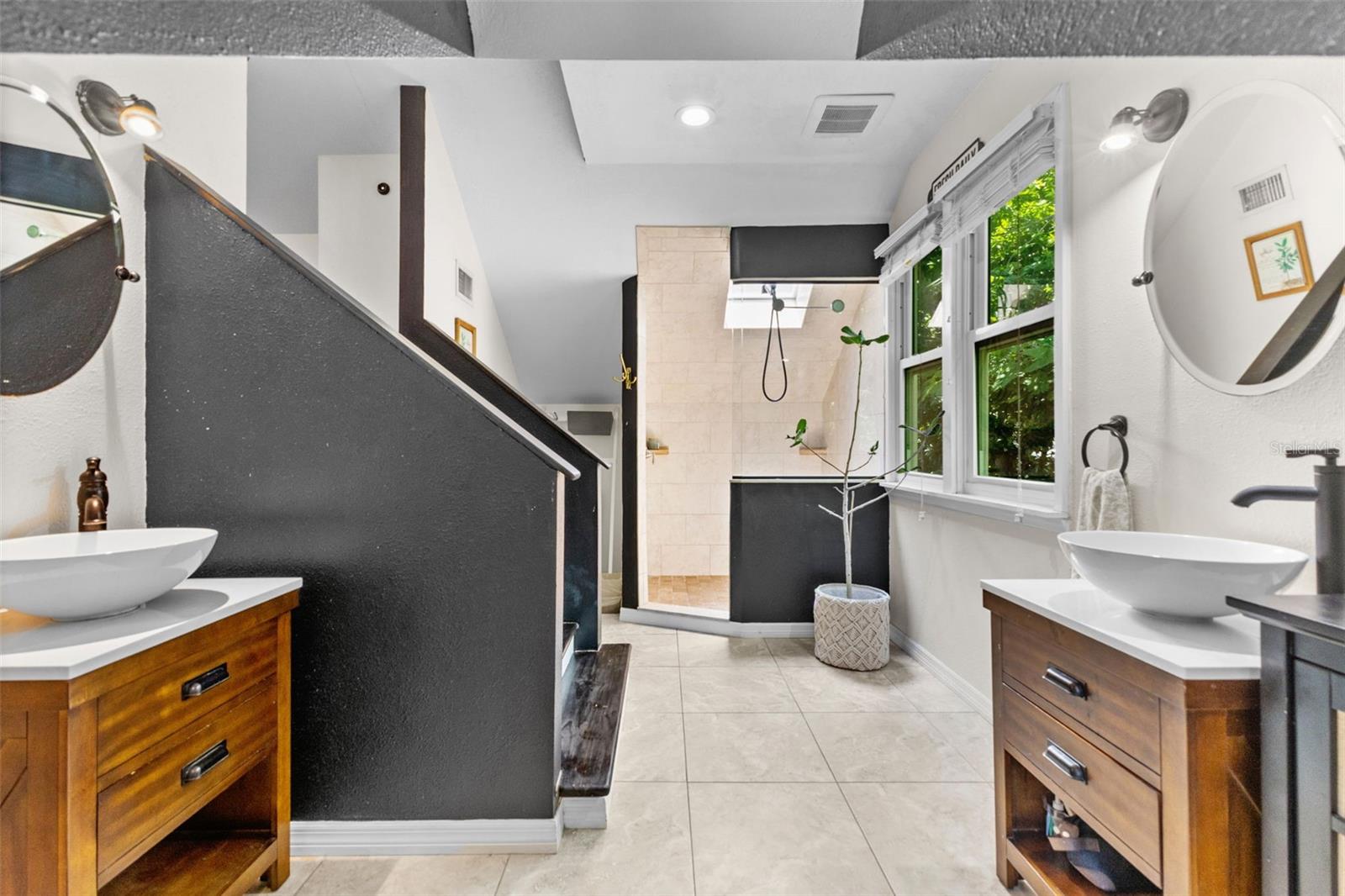 Bright en-suite bath with walk-in tiled shower, skylight, vessel-sink vanity, and a leafy treetop window view.