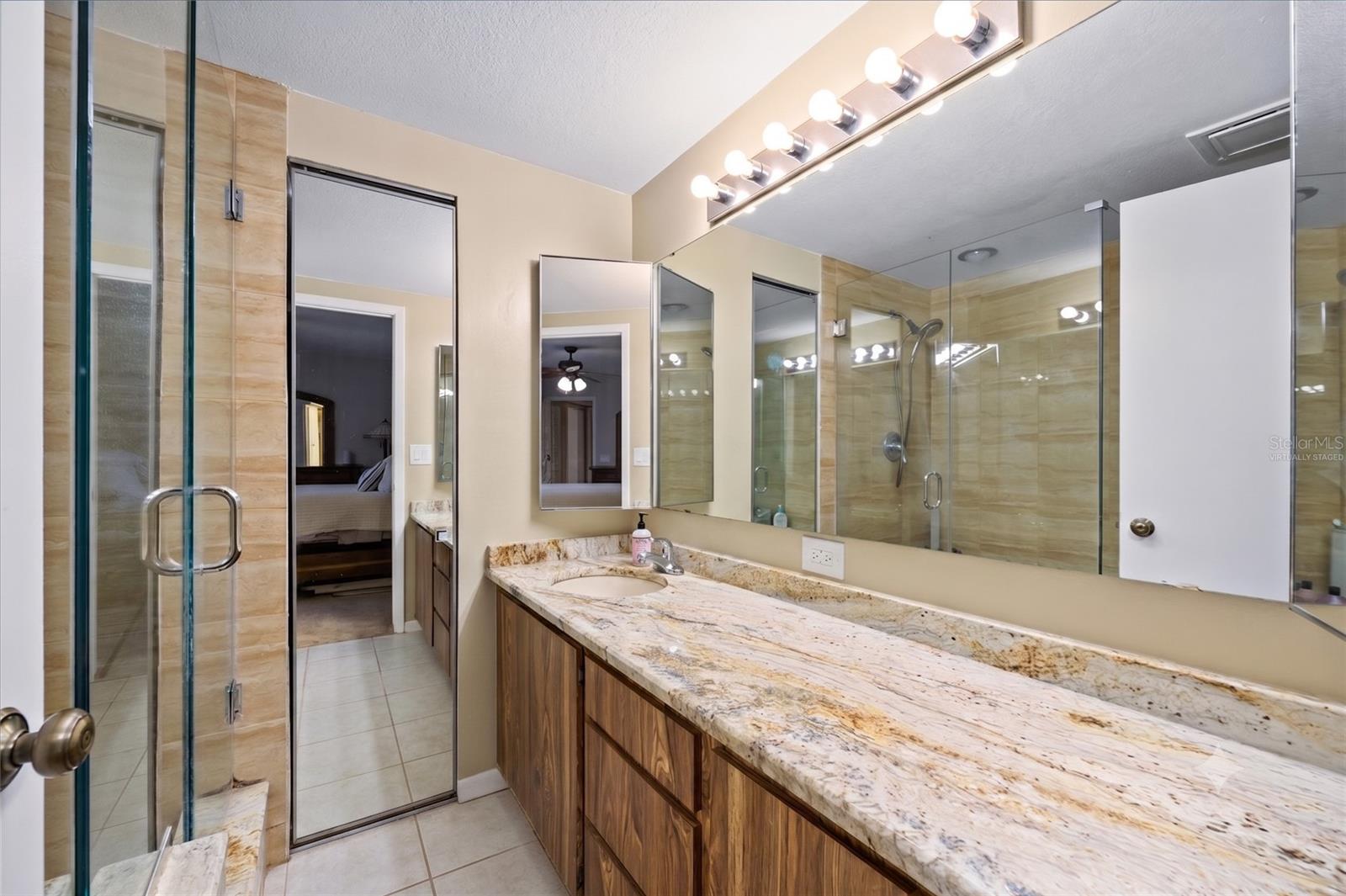 The primary suite bathroom offers a walk-in shower with glass doors, a linen closet, and stunning stone counters. The virtually staged image shows the timeless beauty of this bathroom.