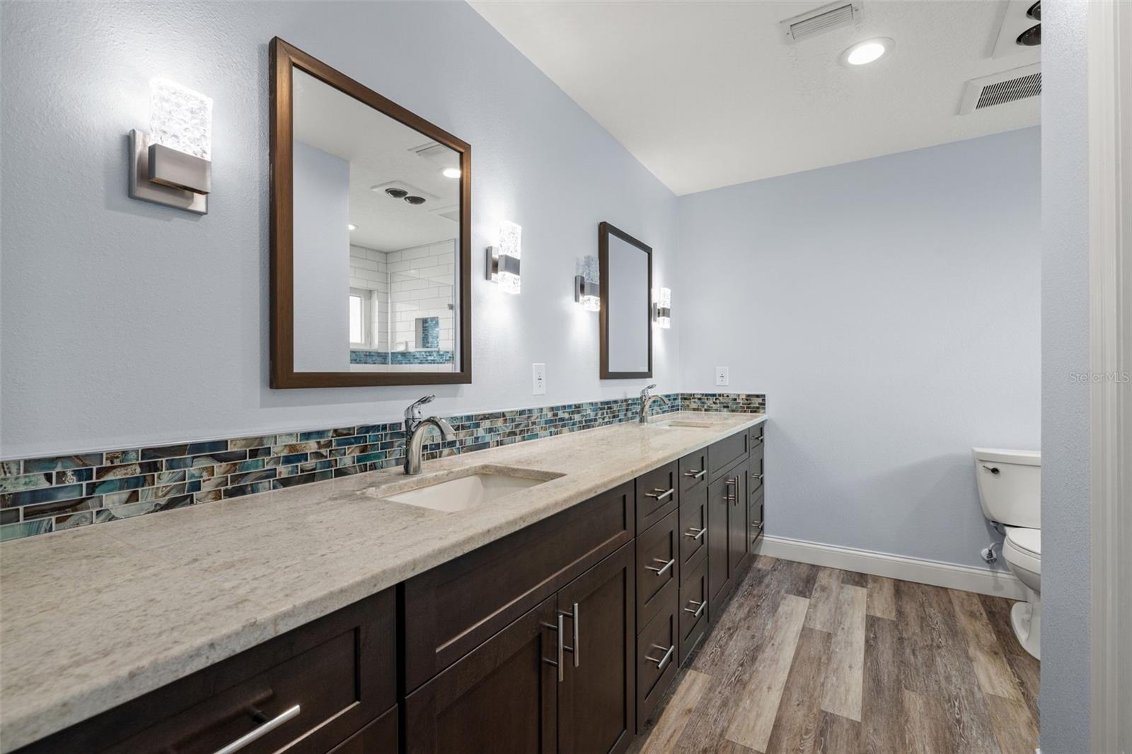A dual-sink vanity in rich espresso-toned cabinetry is finished with a glass mosaic backsplash and stone countertops, giving this renovated primary bath a spa-like polish.