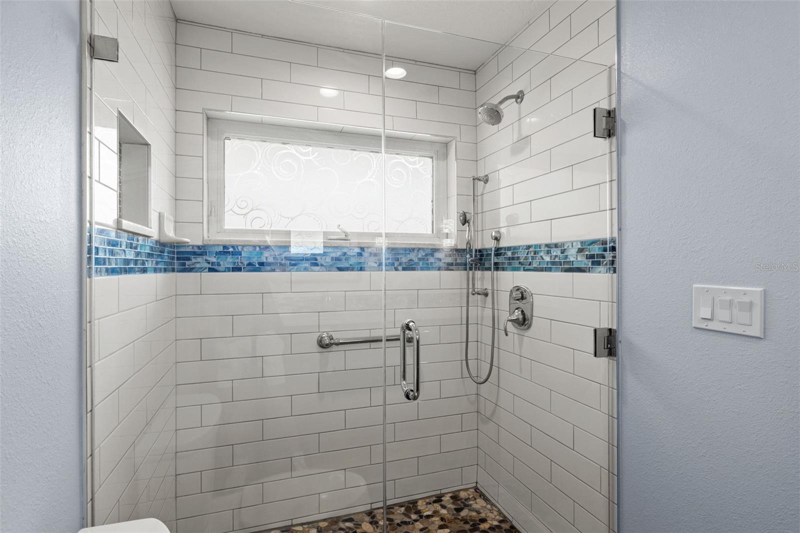 A walk-in shower finished in subway tile with a blue mosaic accent band adds a designer touch to this updated bath, with a window bringing in natural light.