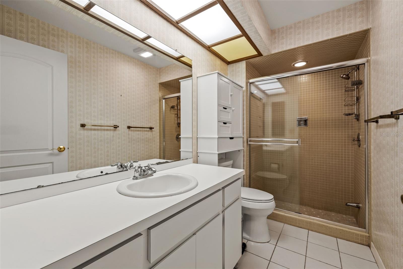 A glass-enclosed, tan-tiled shower pairs with a clean white vanity in this full bathroom, offering a bright, functional layout for family or guests.