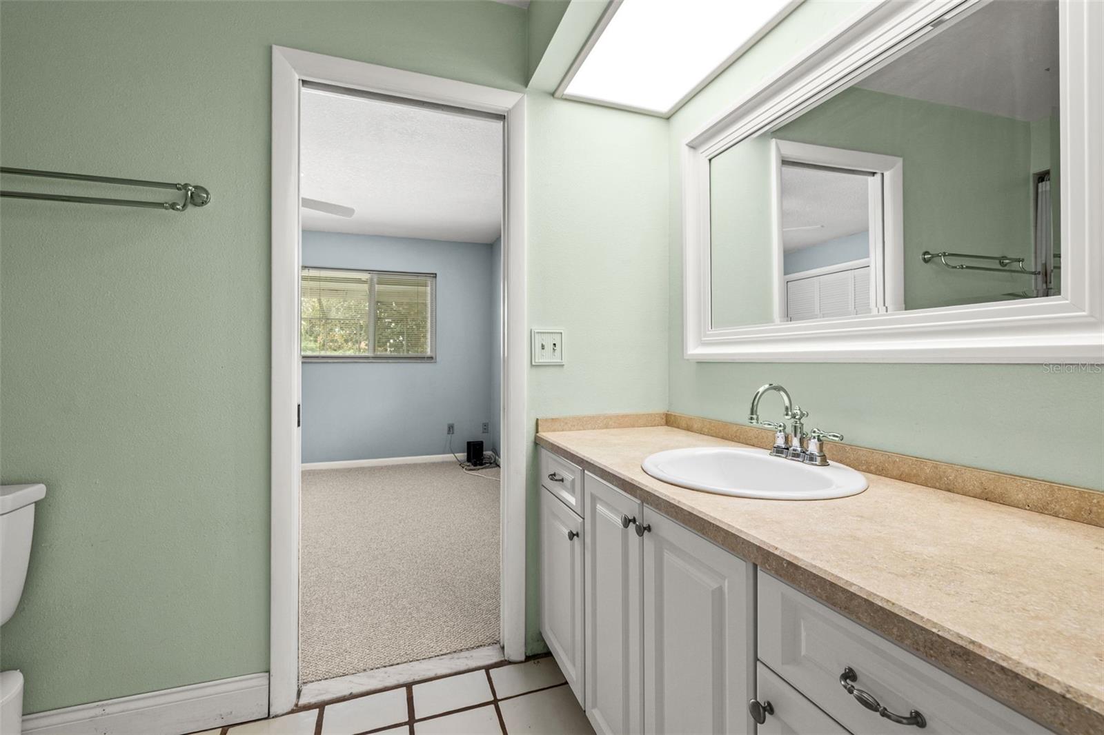 Soft green walls set a calm tone in this Jack and Jill bathroom, with a doorway view leading to a tiled vanity and mirror just beyond.