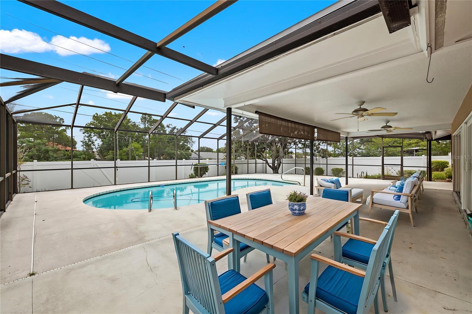 A shaded dining area overlooks the pool, furnished with a wood table and blue cushioned seating that makes indoor-outdoor entertaining effortless. Ceiling fans keep the air moving through the screened lanai, extending everyday living well beyond the back door.