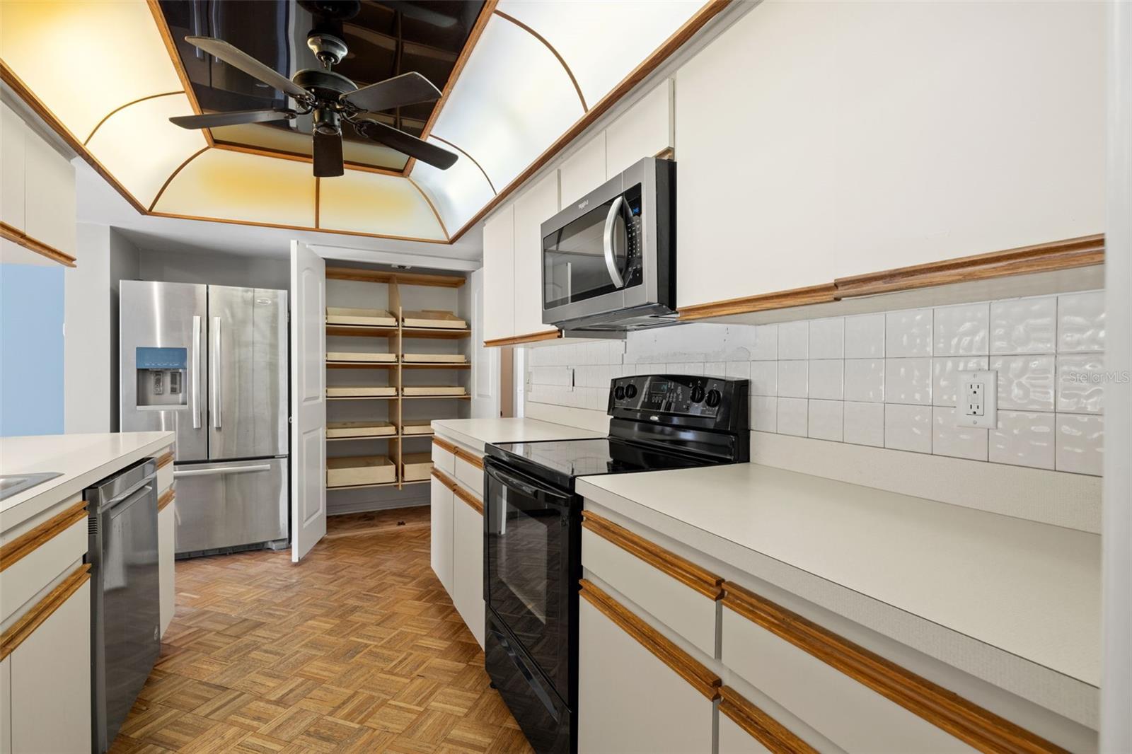 A stainless steel refrigerator and open pantry shelving sit just off the main cooking area, with a domed skylight and ceiling fan overhead keeping the space bright and comfortable.
