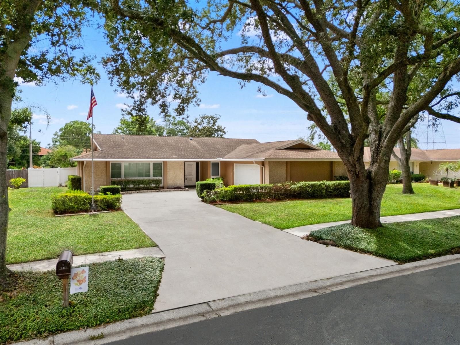 Towering oak trees shade the driveway and front lawn, with irrigated, well-maintained landscaping framing the approach to the home.