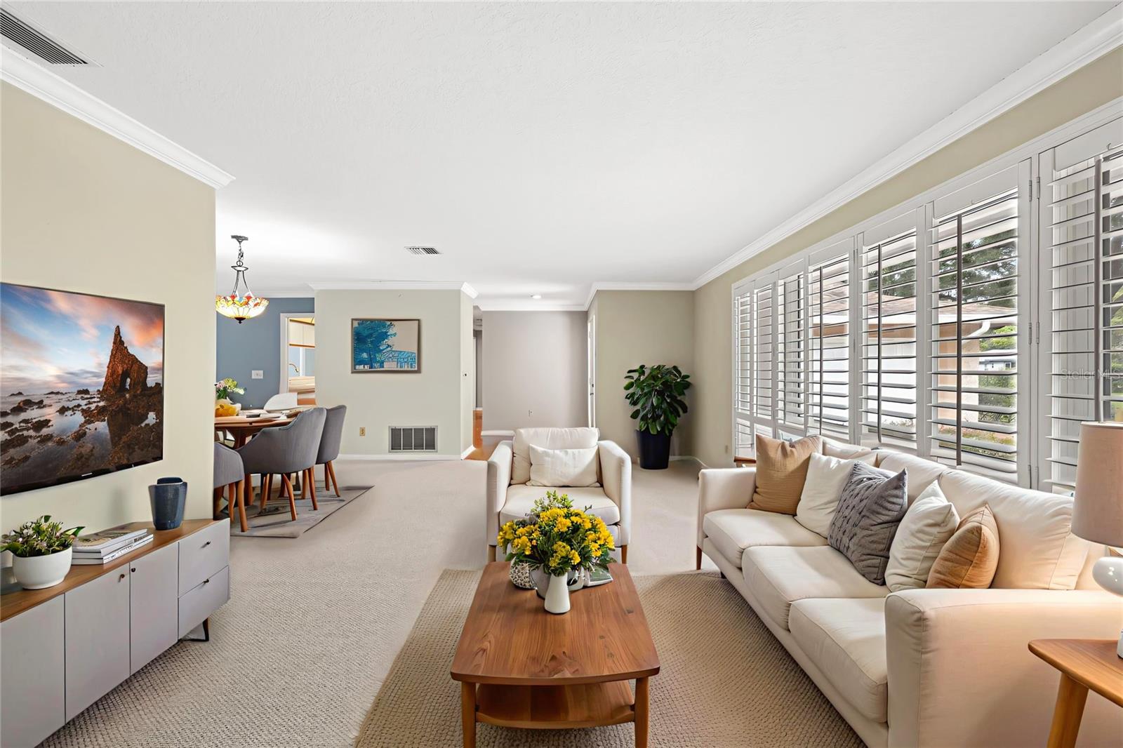 Plantation shutters dress the windows in this sunlit gathering space, softening the light while framing views of the front yard. Crown molding and a cohesive neutral palette carry through the open living and dining area.