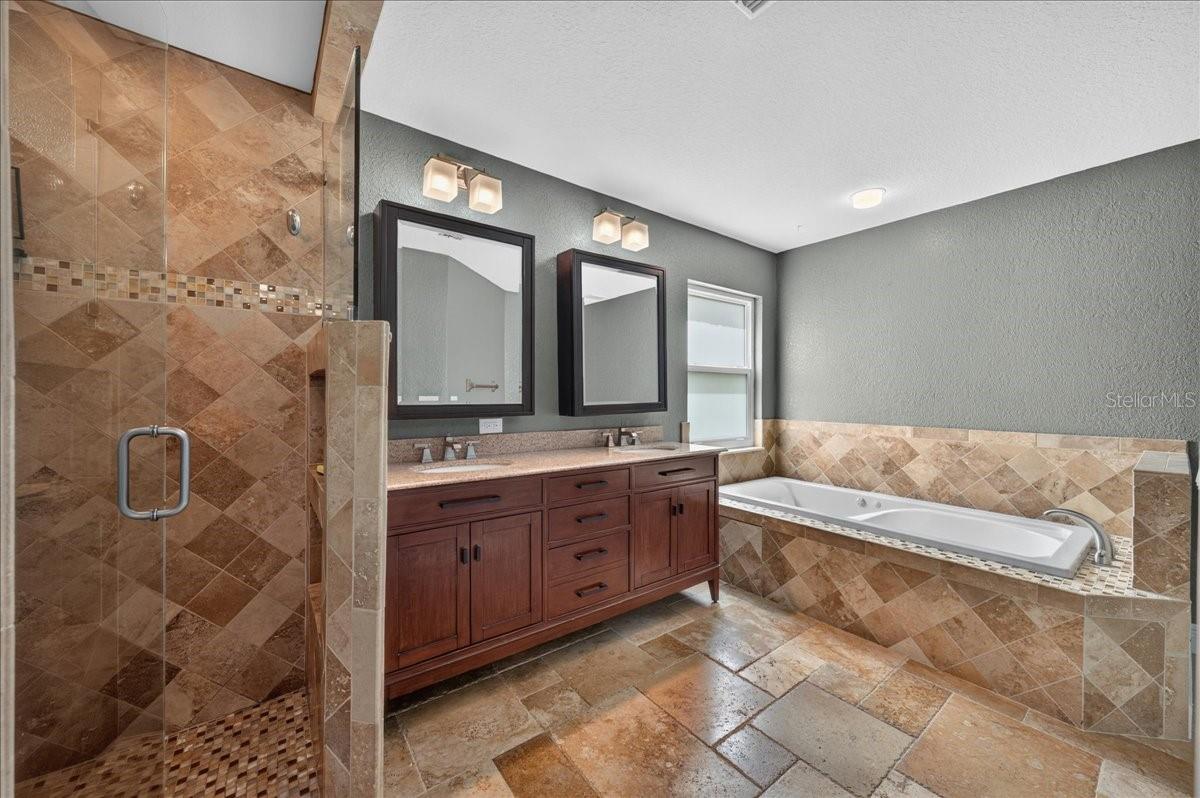 The ensuite bath features travertine flooring, a dual-sink granite vanity, jetted tub, separate walk-in shower, and built-in cabinetry for linens and additional storage.