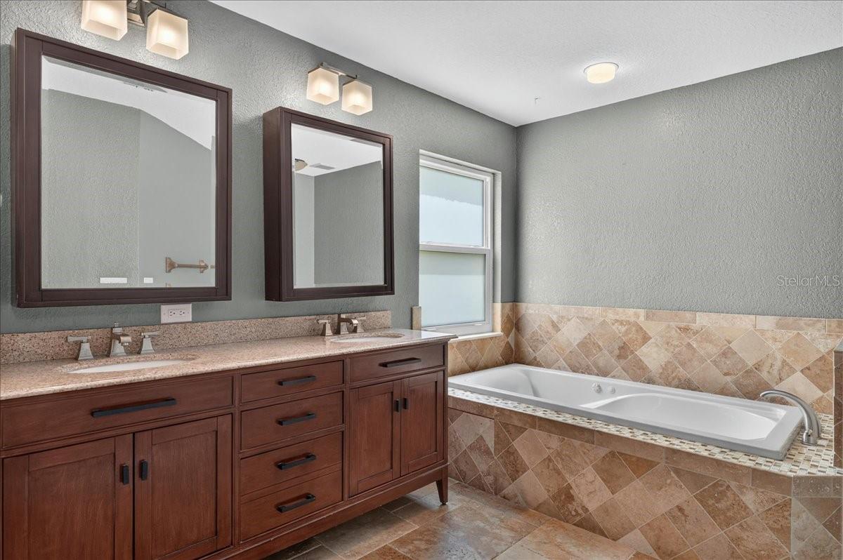 The ensuite bath features travertine flooring, a dual-sink granite vanity, jetted tub, separate walk-in shower, and built-in cabinetry for linens and additional storage.
